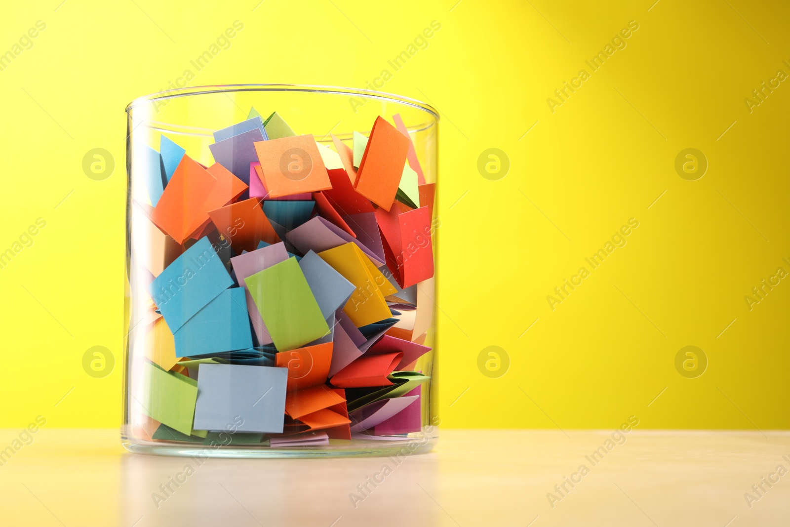 Jar with colorful pieces of paper on wooden table against yellow background. Space for text Photo of Jar with colorful pieces of paper on wooden table against yellow background. Space for text