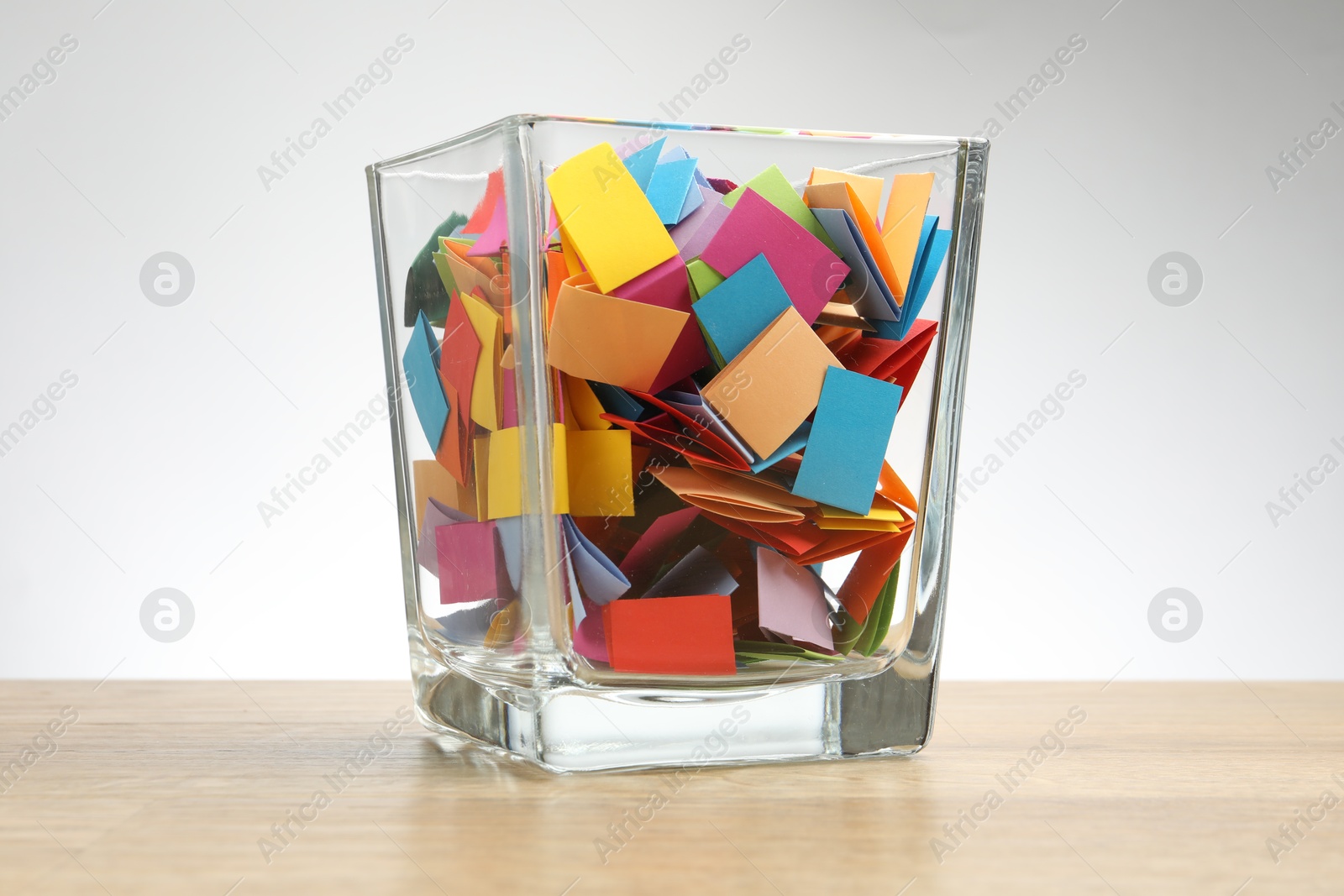 Jar with colorful pieces of paper on wooden table against light background Photo of Jar with colorful pieces of paper on wooden table against light background