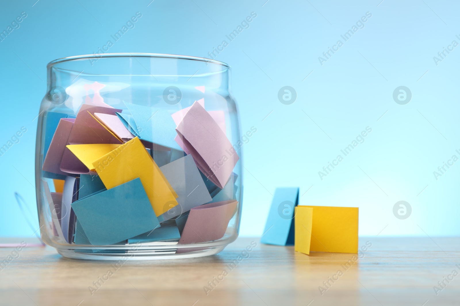 Jar with colorful pieces of paper on wooden table against light blue background. Space for text Photo of Jar with colorful pieces of paper on wooden table against light blue background. Space for text