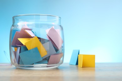 Photo of Jar with colorful pieces of paper on wooden table against light blue background. Space for text
