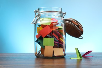 Photo of Jar with colorful pieces of paper on wooden table against light blue background