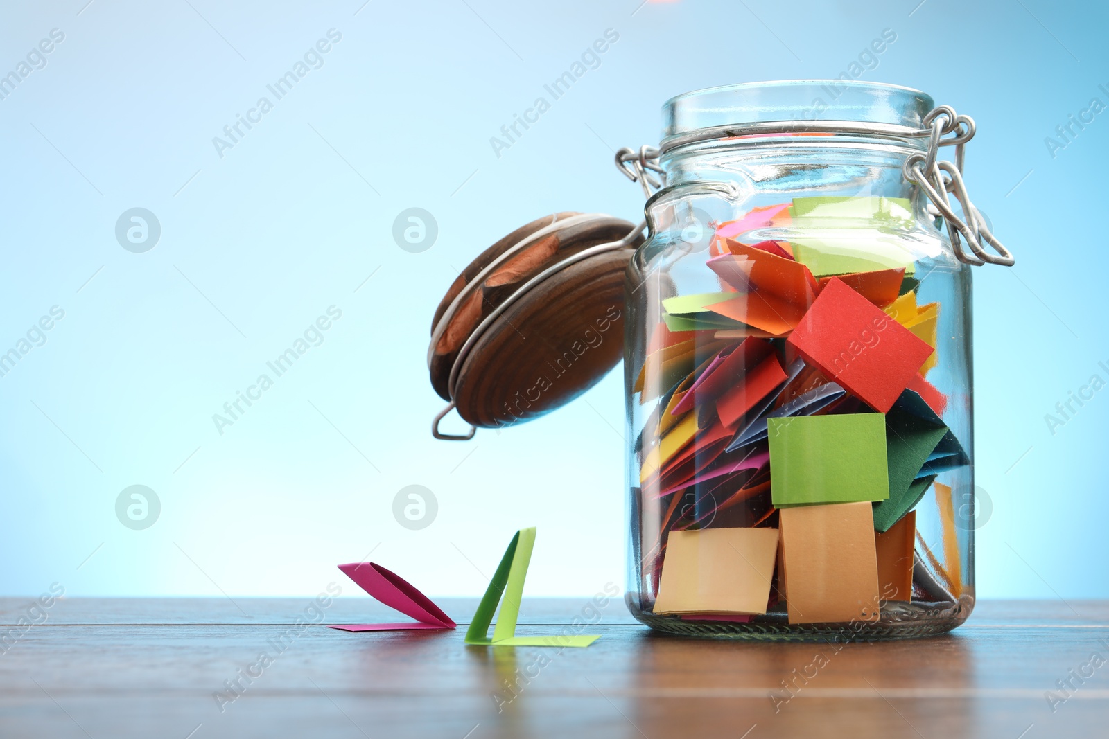 Jar with colorful pieces of paper on wooden table against light blue background. Space for text Photo of Jar with colorful pieces of paper on wooden table against light blue background. Space for text