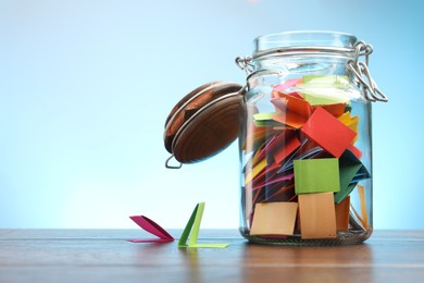 Photo of Jar with colorful pieces of paper on wooden table against light blue background. Space for text