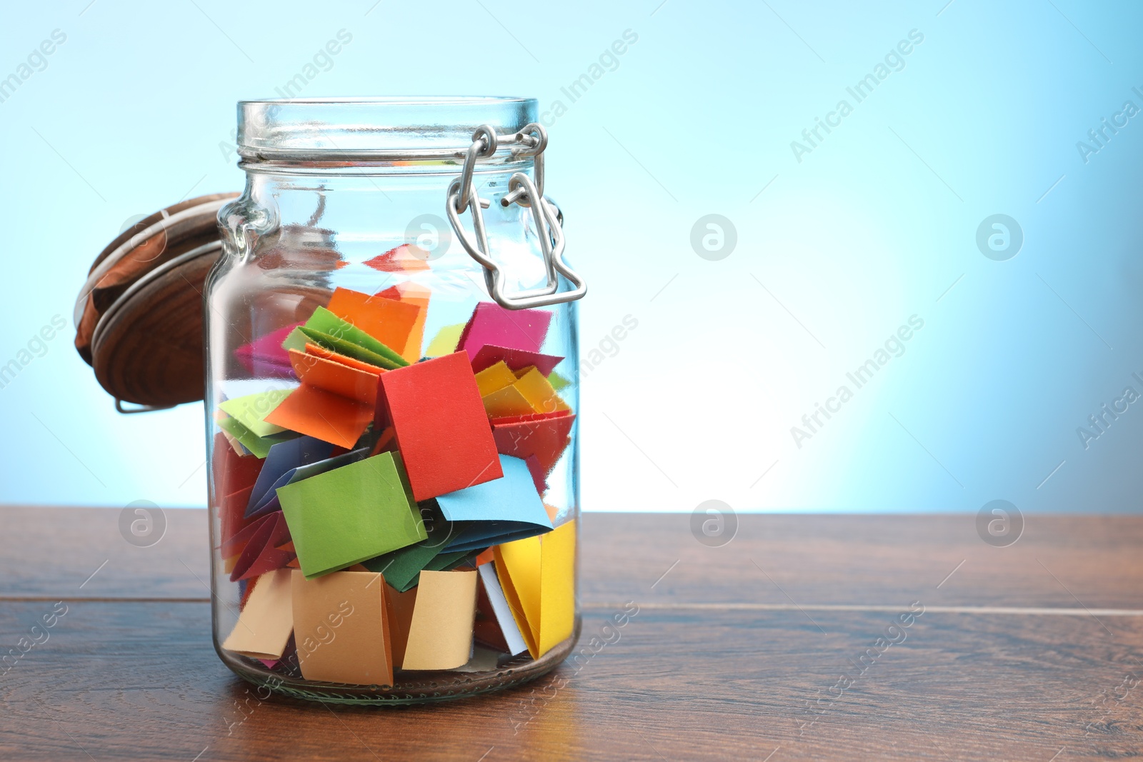 Jar with colorful pieces of paper on wooden table against light blue background. Space for text Photo of Jar with colorful pieces of paper on wooden table against light blue background. Space for text