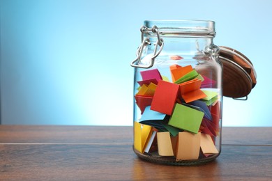Photo of Jar with colorful pieces of paper on wooden table against light blue background. Space for text
