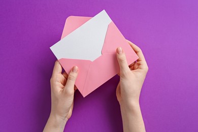 Photo of Woman holding envelope with blank invitation card on purple background, top view
