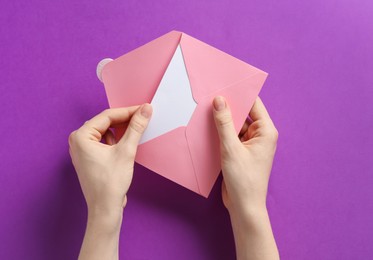 Photo of Woman holding envelope with blank invitation card on purple background, top view