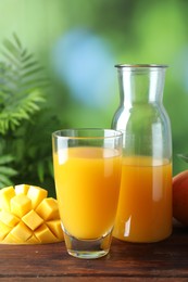Photo of Tasty mango juice in glass, carafe and fresh fruit on wooden table against blurred background