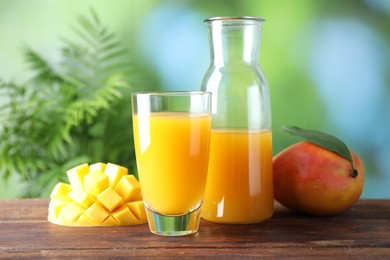 Photo of Tasty mango juice in glass, carafe and fresh fruits on wooden table against blurred background