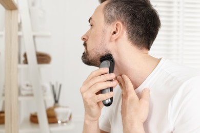 Photo of Handsome man shaving with trimmer near mirror at home