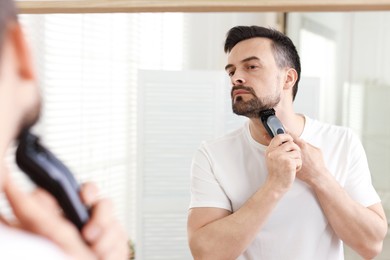 Photo of Handsome man shaving with trimmer near mirror at home