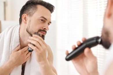 Photo of Handsome man shaving with trimmer near mirror at home