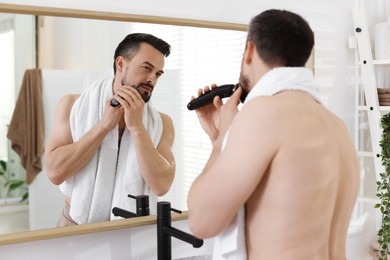 Photo of Handsome man shaving with trimmer near mirror at home
