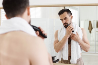 Photo of Handsome man shaving with trimmer near mirror in bathroom