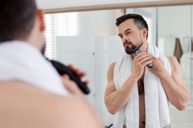 Photo of Handsome man shaving with trimmer near mirror in bathroom