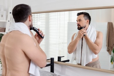 Photo of Handsome man shaving with trimmer near mirror in bathroom