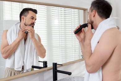 Photo of Handsome man shaving with trimmer near mirror in bathroom