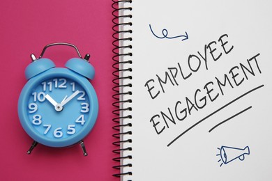 Image of Employee engagement. Words and drawings in notebook and alarm clock on pink background, top view