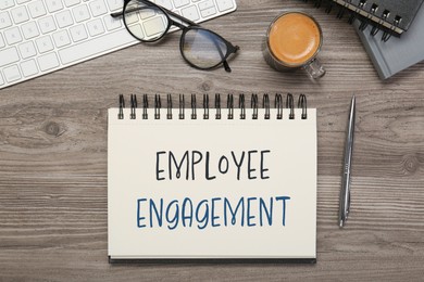 Image of Employee engagement. Words in notebook, pen and computer keyboard on wooden table, top view