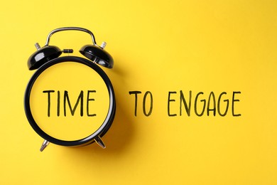 Image of Time to engage. Black alarm clock and words on yellow background, top view