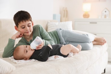Photo of Little boy feeding his newborn brother with bottle on sofa at home