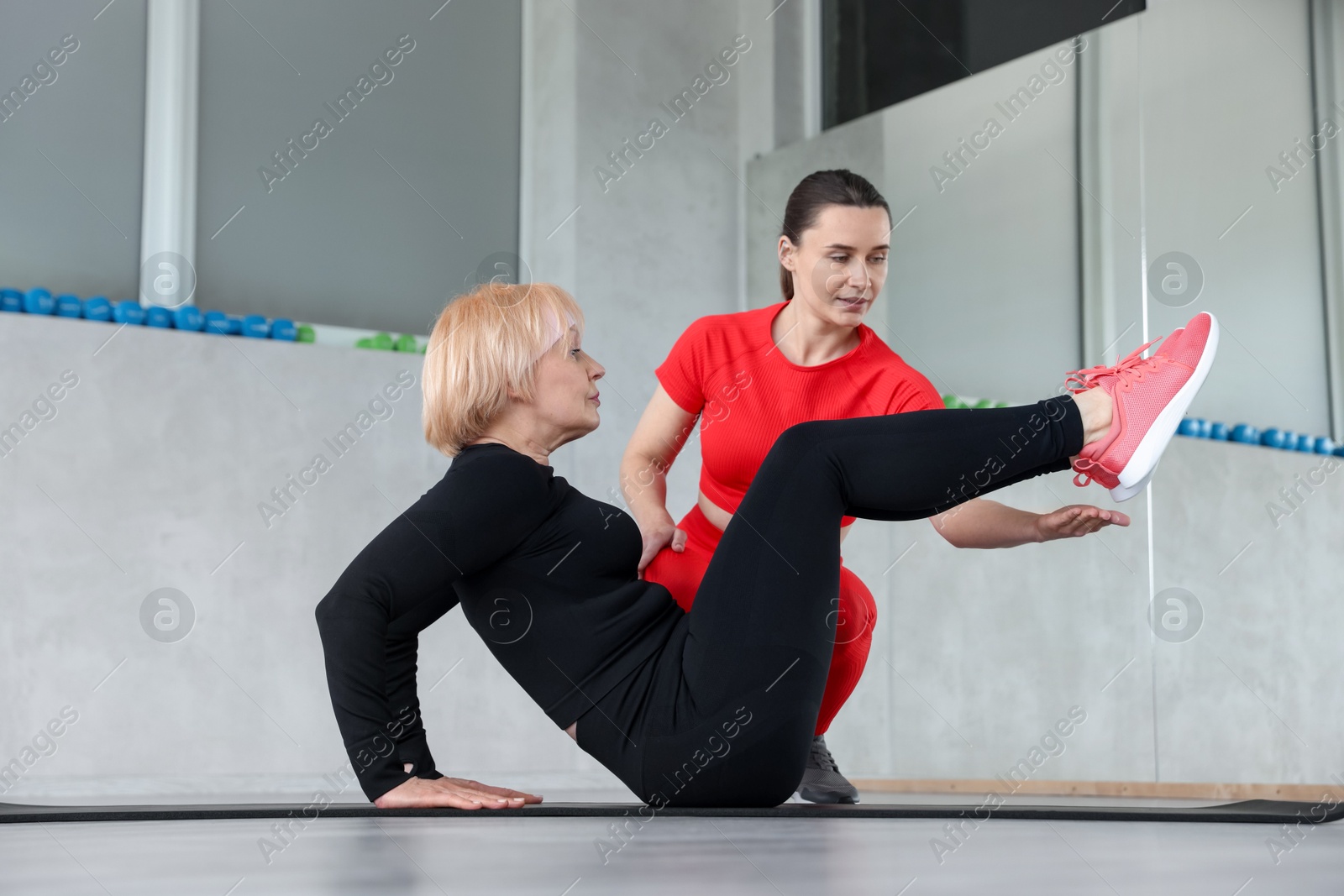 Senior woman exercising on yoga mat supervised by professional trainer in gym Photo of Senior woman exercising on yoga mat supervised by professional trainer in gym