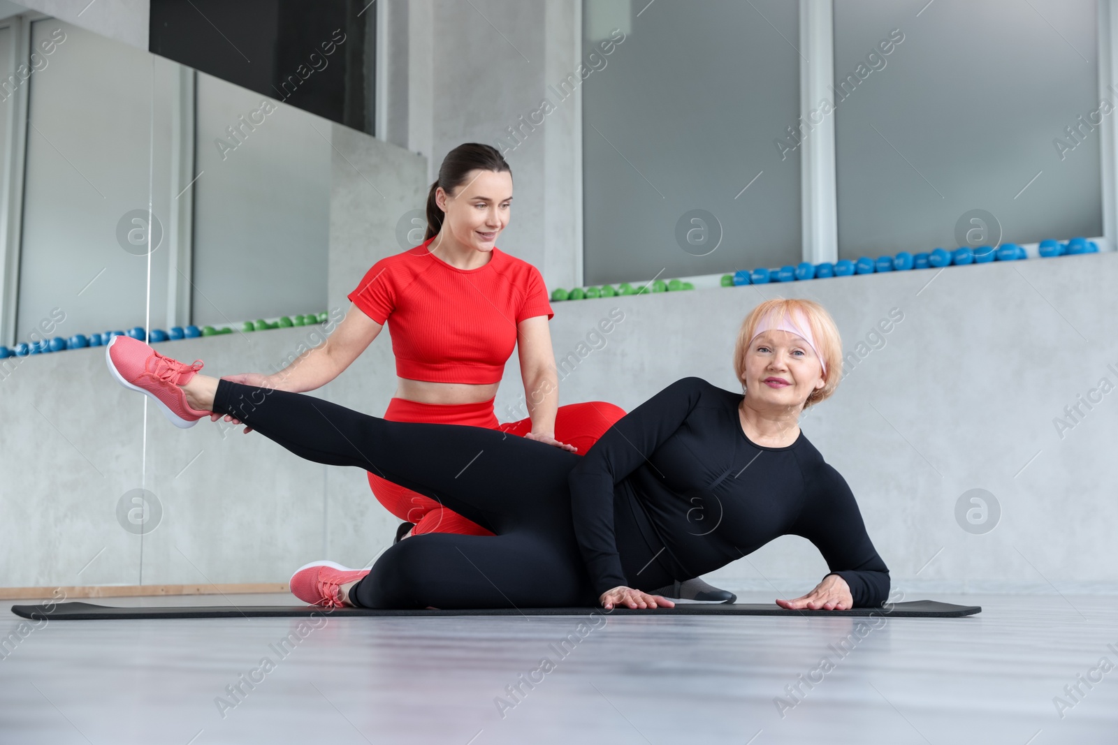 Senior woman exercising on yoga mat supervised by professional trainer in gym Photo of Senior woman exercising on yoga mat supervised by professional trainer in gym