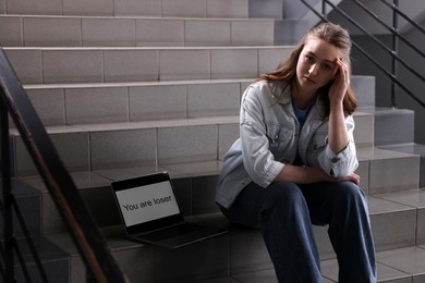 Photo of Cyber bullying. Upset teenage girl and laptop with text You Loser on stairs indoors