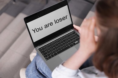 Photo of Cyber bullying. Upset teenage girl looking at laptop with text You Loser on stairs indoors, closeup
