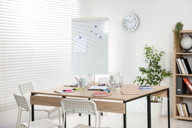 Photo of Wooden table with stationery, chairs and whiteboard in office. Interior design