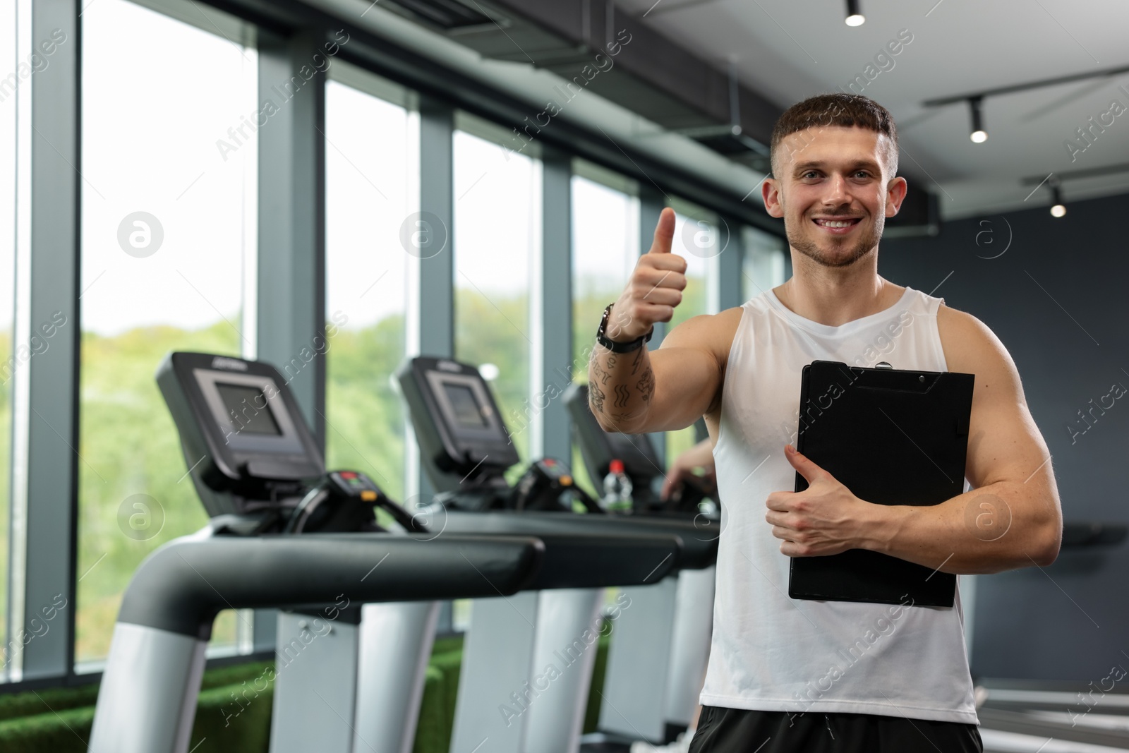 Photo of Personal trainer with clipboard showing thumbs up in sport center. Space for text