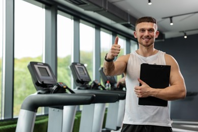 Photo of Personal trainer with clipboard showing thumbs up in sport center. Space for text