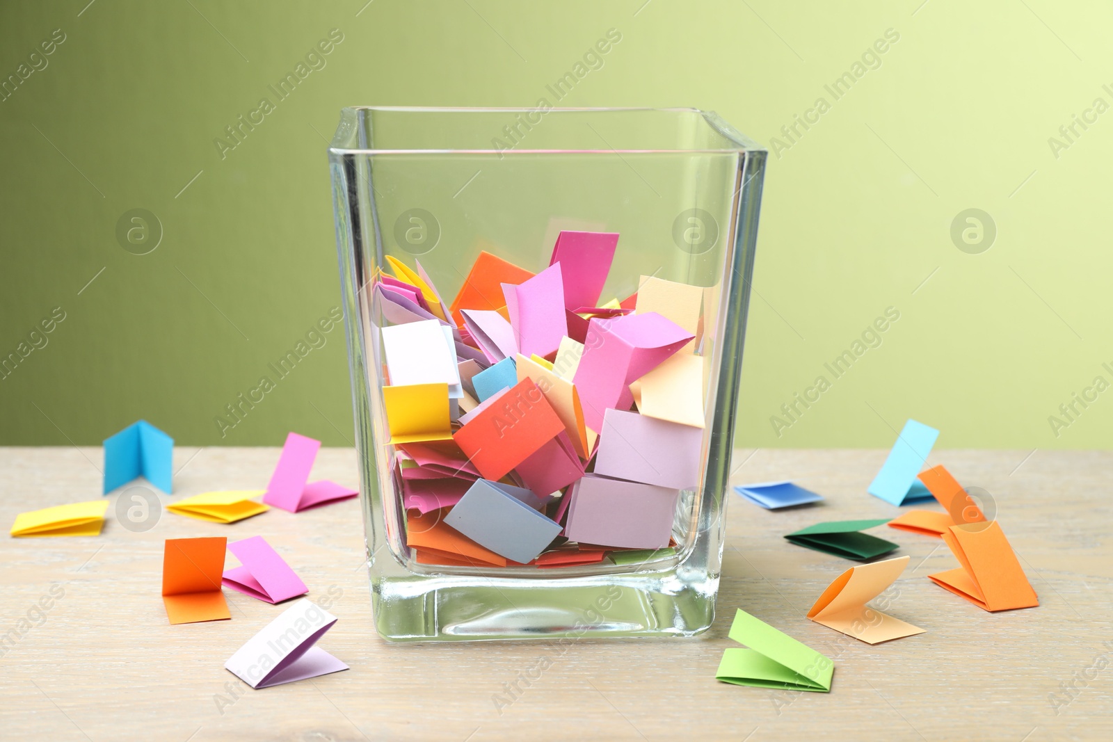 Photo of Jar with colorful pieces of paper on wooden table against green background, closeup