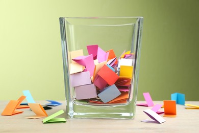 Photo of Jar with colorful pieces of paper on wooden table against green background, closeup