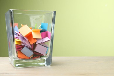 Photo of Jar with colorful pieces of paper on wooden table against green background, closeup. Space for text