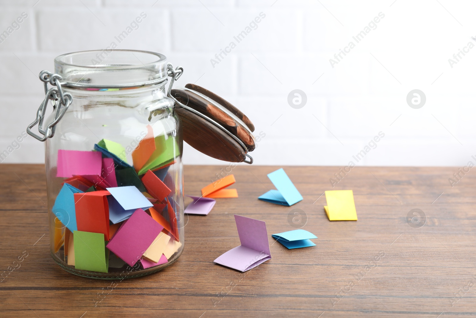 Jar with colorful pieces of paper on wooden table near white brick wall. Space for text Photo of Jar with colorful pieces of paper on wooden table near white brick wall. Space for text
