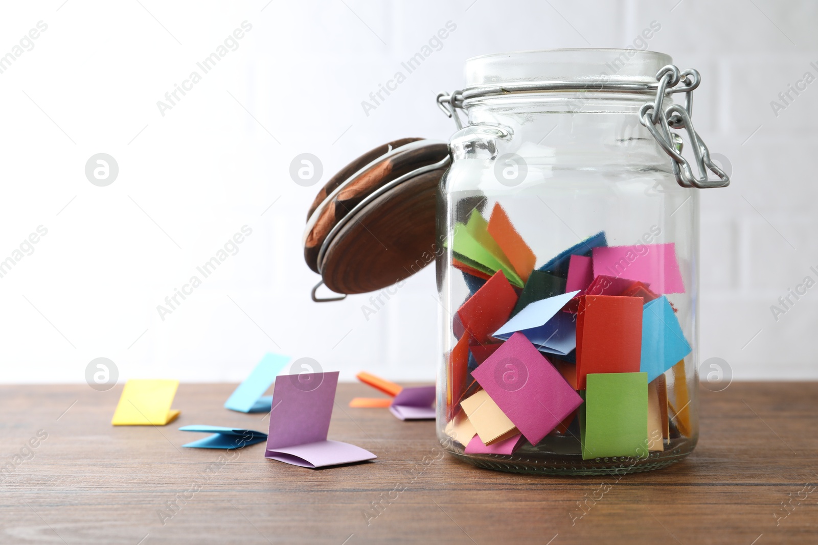 Jar with colorful pieces of paper on wooden table near white brick wall. Space for text Photo of Jar with colorful pieces of paper on wooden table near white brick wall. Space for text