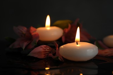 Photo of Burning candles and beautiful flowers on wet black table, closeup