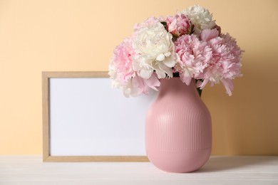 Photo of Beautiful peonies in vase and picture frame on white table against beige background