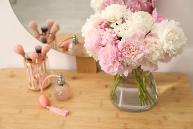 Photo of Beautiful peonies in vase, perfume, brushes and mirror on wooden vanity indoors