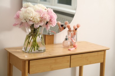 Photo of Beautiful peonies in vase, perfume, brushes and mirror on wooden vanity indoors