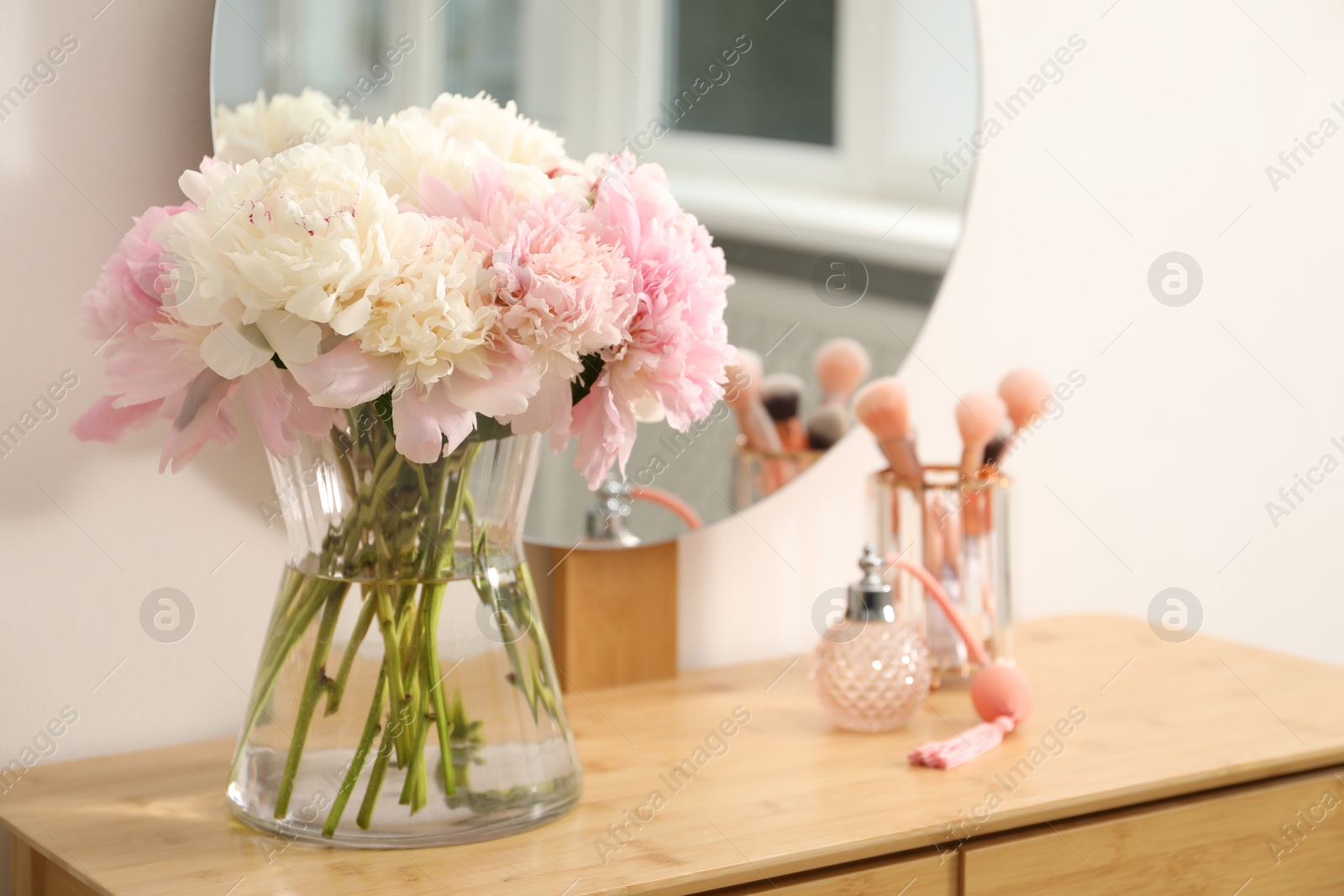 Beautiful peonies in vase, perfume, brushes and mirror on wooden vanity indoors Photo of Beautiful peonies in vase, perfume, brushes and mirror on wooden vanity indoors