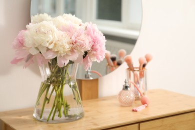 Photo of Beautiful peonies in vase, perfume, brushes and mirror on wooden vanity indoors