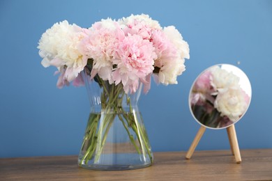 Photo of Beautiful peonies in vase and mirror on wooden table near light blue wall