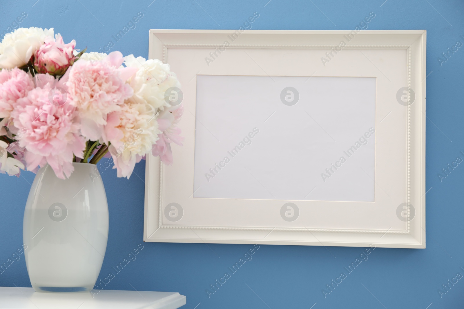Beautiful peonies in vase and picture frame on white table near light blue wall Photo of Beautiful peonies in vase and picture frame on white table near light blue wall
