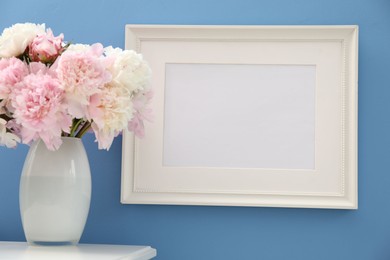 Photo of Beautiful peonies in vase and picture frame on white table near light blue wall