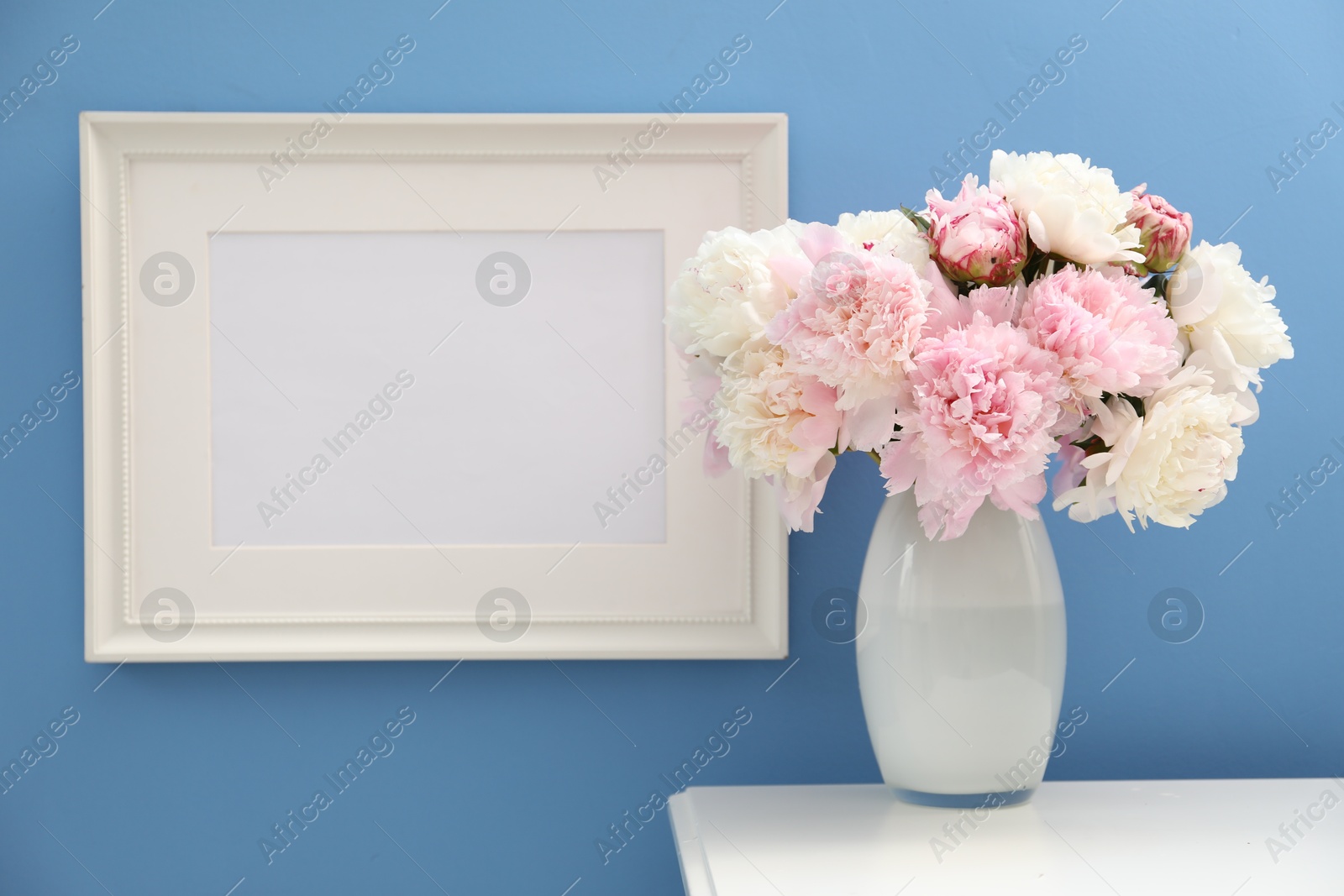 Beautiful peonies in vase and picture frame on white table near light blue wall Photo of Beautiful peonies in vase and picture frame on white table near light blue wall