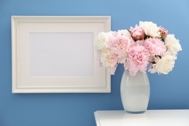 Photo of Beautiful peonies in vase and picture frame on white table near light blue wall