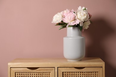 Photo of Beautiful peonies in vase on wooden dresser near pink wall. Space for text