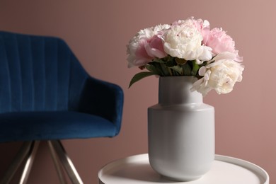 Photo of Beautiful peonies in vase on white table and armchair near pink wall